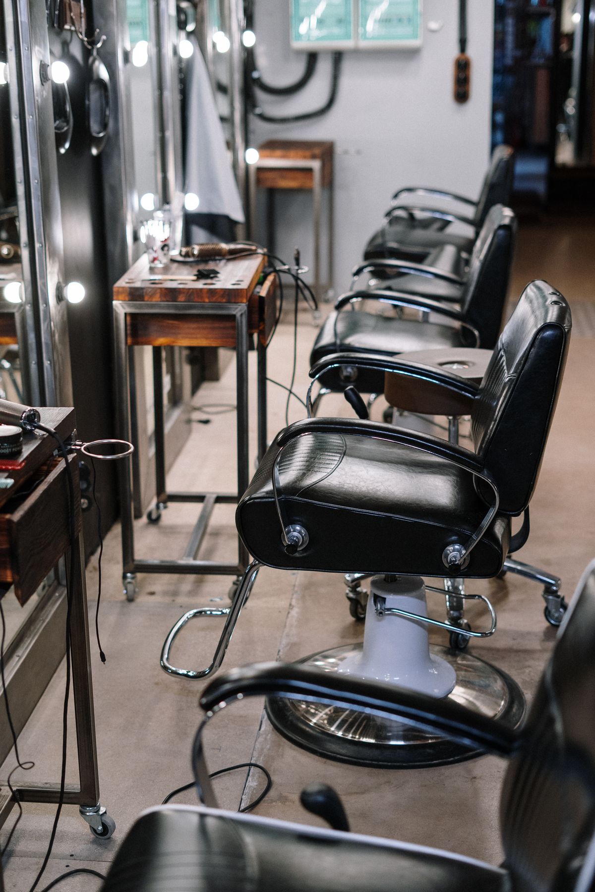A row of black leather barber chairs beside a wall of bulb-lit mirrors, with wooden trolleys holding clippers and combs on the left.