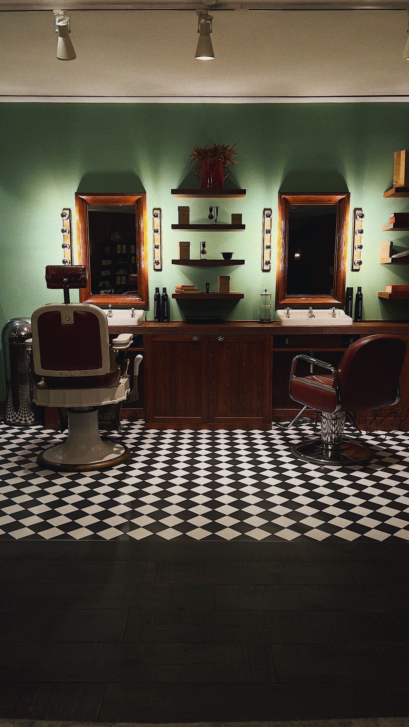 Interior of a vintage-style barbershop: a green wall with two wood-framed mirrors with bulb lights, floating shelves holding grooming bottles and a plant, two basins, a white-and-red vintage barber chair and a red leather chair on a black-and-white checkerboard floor.