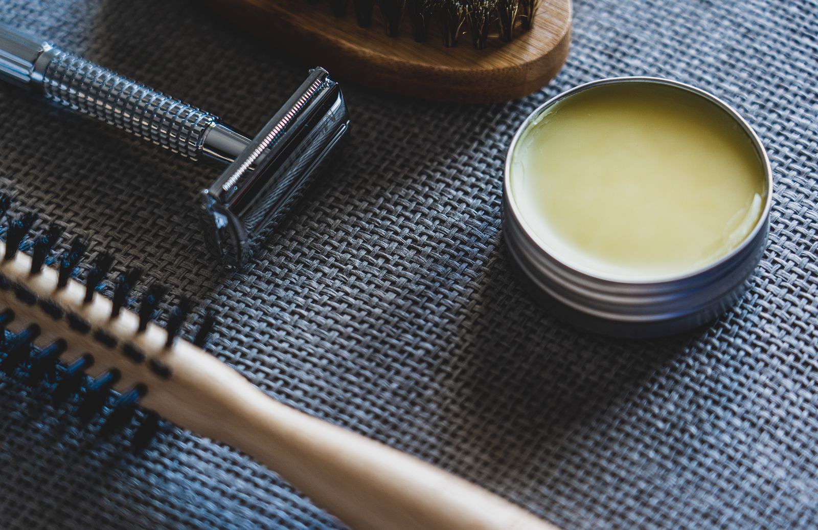 A close-up on a grey woven mat of a chrome safety razor, a round tin of pale-yellow balm, a bristle brush at top and a round wooden hair brush at bottom left.