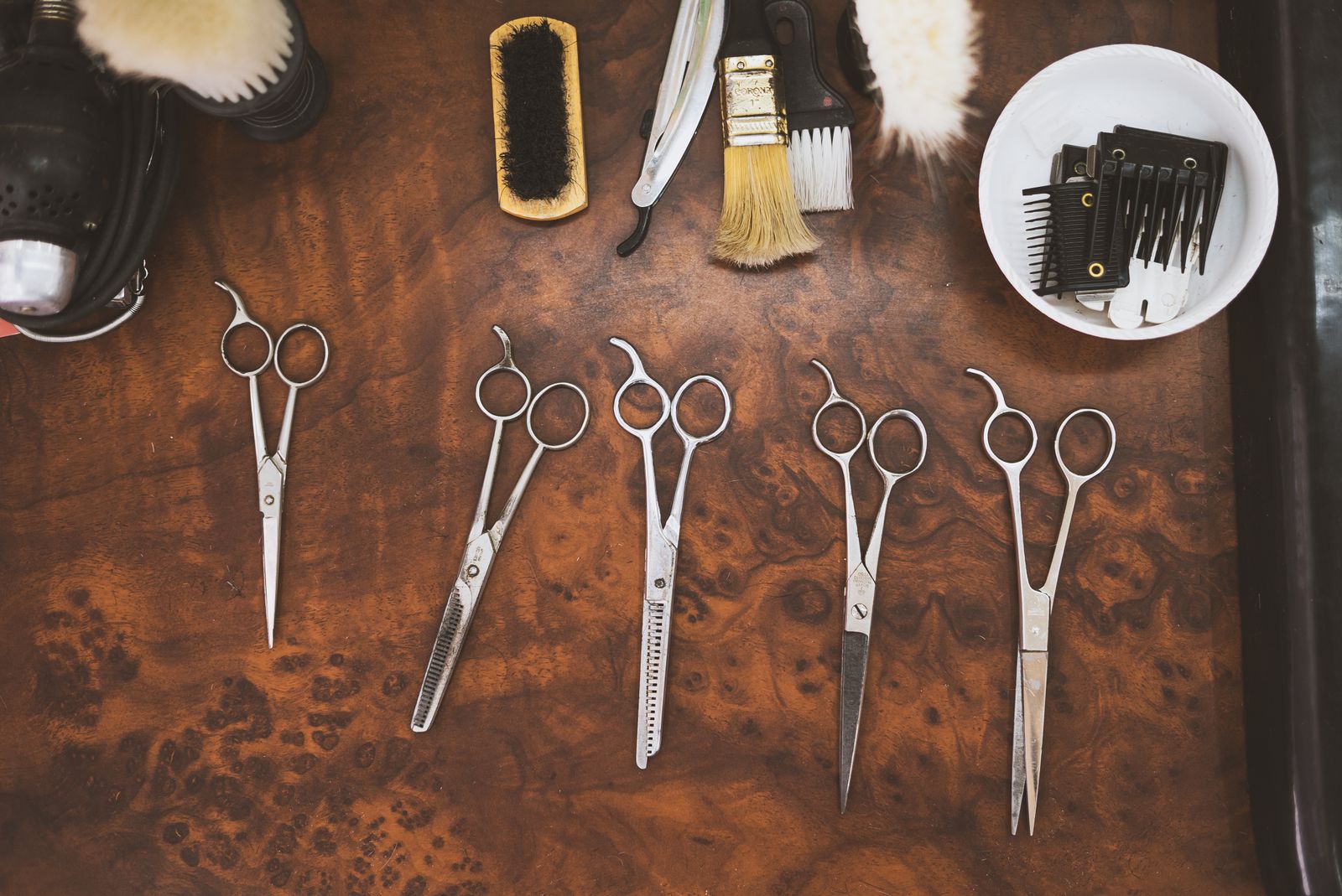 An overhead view of five pairs of steel scissors including thinning shears laid out on a burr-wood surface, with brushes, a razor handle and a bowl of black clipper guard combs along the top.